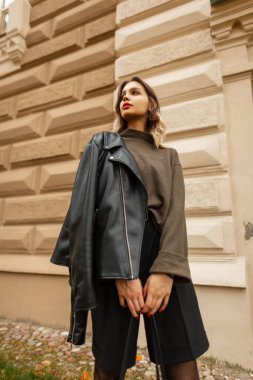 Moda kıyafetleri içinde, yeşil kazaklı, siyah deri ceketli ve şortlu güzel bir kadın sonbahar günü eski bir binanın yanında duruyor.