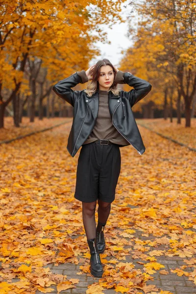 黒革のジャケット 緑のセーター ショートパンツ 爪と葉と明るい黄金の秋の公園を歩く靴とファッショナブルな秋の服でスタイリッシュな若い女の子 — ストック写真