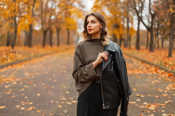 一个穿着时尚绿色毛衣和黑色皮夹克 红唇的年轻貌美女子的秋天女服务员走在一个金黄色的秋天公园里 — 图库照片