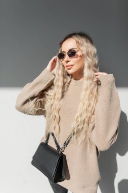 Gri bir arkaplanda güneşli bir günde, güzel, modaya uygun, kıvırcık saçlı, çantalı ve klasik güneş gözlüklü, güzel, güzel, şık sarışın bir kadın.