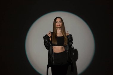 Moda kıyafetleri içinde güzel bir kız. Üstü açık, siyah bir arka planda deri ceketi var.