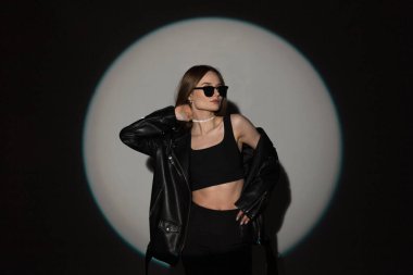 Moda stüdyosu genç bir kız portresi. Moda güneş gözlüklü, şık rock kıyafetleri içinde. Üstü açık, koyu renk arka planda deri bir ceket.