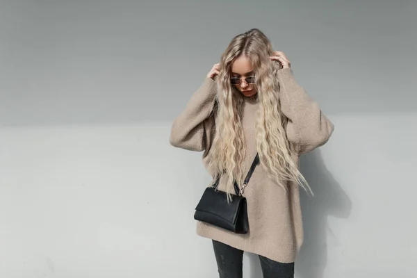 スタイリッシュな若い女性とブロンドの巻き毛を持つヴィンテージサングラスでベージュのセーターと流行の黒のバッグの近くに灰色の壁通り — ストック写真