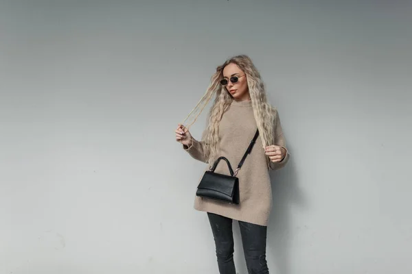 ファッショナブルな美しい都市の女性モデルとともに巻きブロンドの髪型ヴィンテージサングラスでスタイリッシュなベージュのセーターでファッション黒のバッグは 市内の灰色の壁の近くに立つ — ストック写真