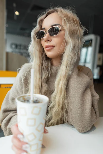 곱슬곱슬 금발의 아름다운 아가씨가 베이지 스웨터를 안경을 종이컵을 카페에 — 스톡 사진