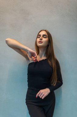 Zarif siyah elbiseli, üstü ve pantolonu gri bir duvarın yanında poz veren moda güzeli bir kadın.
