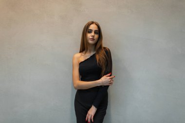 Şık bir bluzu ve pantolonu olan şık siyah elbiseli güzel moda kadını gri bir duvarın yanında duruyor.