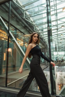 Moda siyah elbiseli, üst ve pantolonu olan şık, genç ve güzel bir işletme yöneticisi modern cam ofis binasında yürüyor. Güzel bir kadın içeride hareket ediyor.