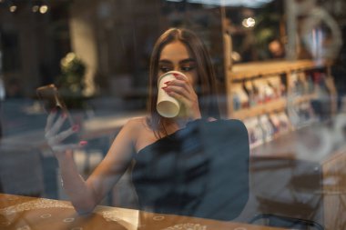 Siyah bluzlu güzel bir model, pencerenin yanındaki kafede oturuyor, kahve içiyor ve akıllı telefondan mesajlar okuyor.