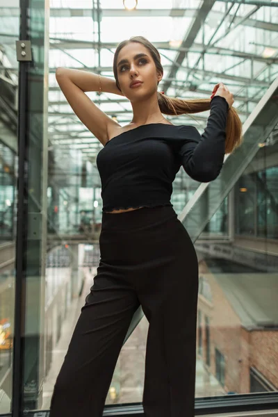 Beautiful young woman with a slender body in a fashion elegant business clothes with a top and trousers stands and fixes her hair in a modern office glass building