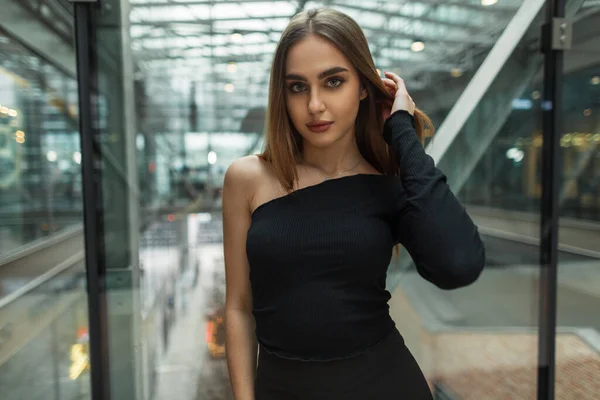 Fashionable beautiful model woman in stylish black clothes with a stylish top stands in a glass office building