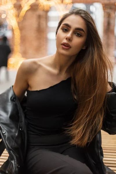 时尚的性感女人穿着时髦的黑色衣服 头戴单肩上衣 身穿羽绒服 有着迷人而新鲜的自然脸蛋和嘴唇 坐在城市里 背景模糊 — 图库照片