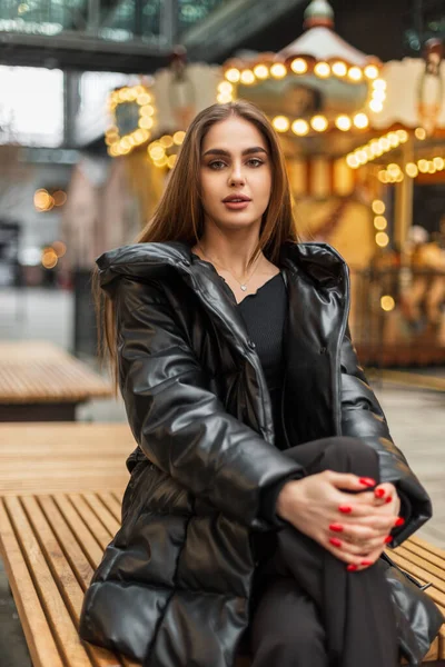 ダウンジャケットとスタイリッシュな黒い冬の服のファッションはかなりエレガントな女性は市内のベンチに座っています — ストック写真