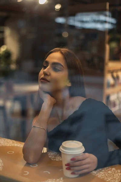 stock image Beautiful young woman model in a fashion top is sitting in a cafe, dreaming and drinking coffee in cafe near the window