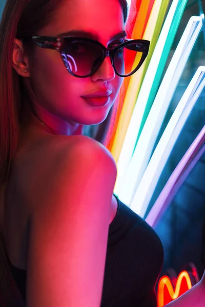 Fashion colorful female portrait of beautiful woman with trendy sunglasses in stylish black dress stands on a dark background with colored neon lights