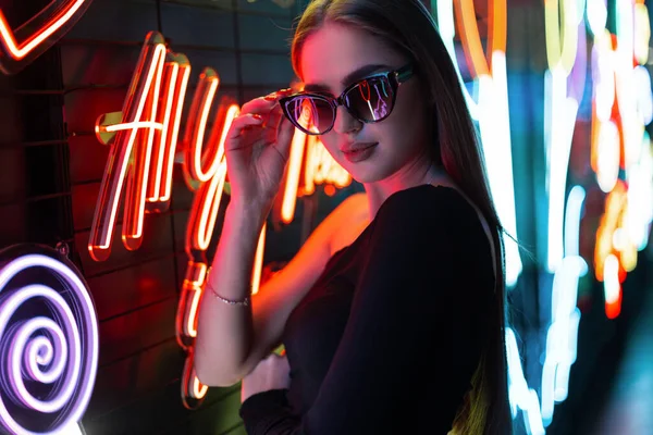 Fashion beautiful cool woman in a black dress wearing cool sunglasses on a dark background of colorful neon sign lights