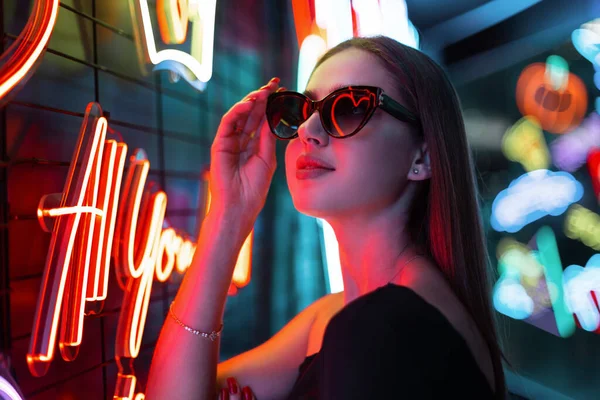 Stylish beautiful night model girl in black fashion dress wears sunglasses and walks on a dark background with neon signs