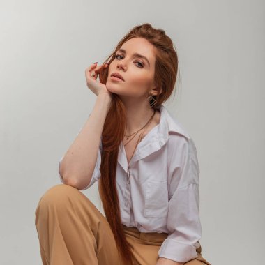 Kızıl saçlı, uzun kızıl saçlı, beyaz tişörtlü, temiz yüzlü güzel bir kadın beyaz arka planda oturuyor.