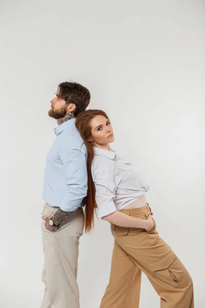 Beautiful young fashion couple man and pretty woman in fashionable casual clothes with shirt, pants standing on a white background in the studio. Redheaded girl and hipster guy standing back to back