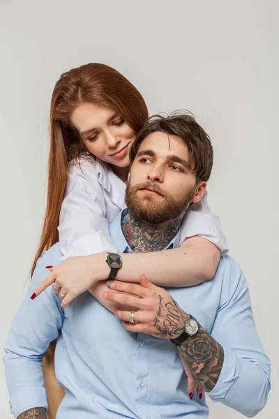 Beautiful young redhead woman in a white shirt hugs a handsome hipster guy with a beard and a tattoo in a blue shirt on a white background. Fashion couple