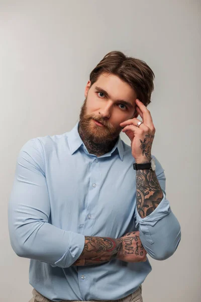 Handsome successful businessman man with a tattoo and a beard in a fashionable blue shirt stands and thinks on a white background in the studio