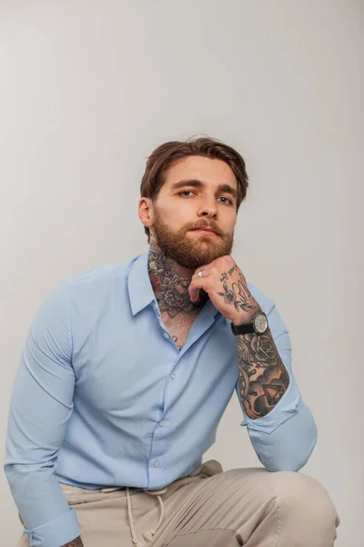 Handsome successful business hipster man with tattoos and beard in fashion blue shirt sits on a white background