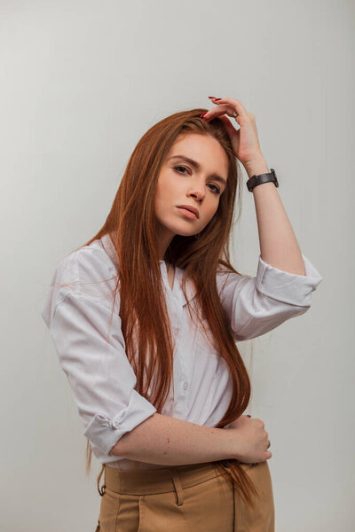 Fashion redhead beauty woman with long red hair in elegant clothes with shirt and pants stands on a white background