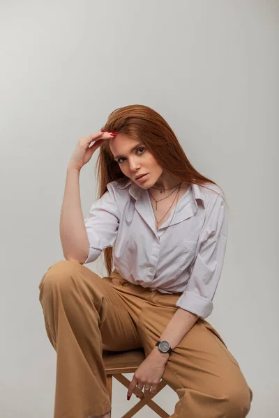 Fashionable beautiful redhead woman model in stylish elegant clothes with shirt and trousers sits on a wooden chair in the studio