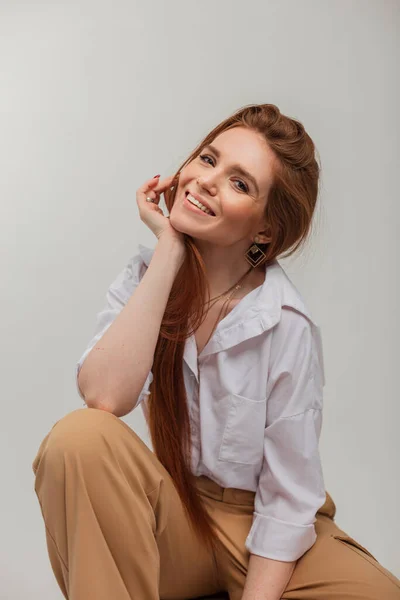 Natural female portrait of a beautiful cheerful cute woman with a beautiful smile in fashion clothes with trousers and a shirt sits on a white background in the studio