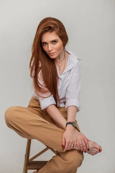 Fashion beautiful fresh redhead woman model with long hair in fashionable elegant clothes with white shirt and pants sits on wooden chair on a white background