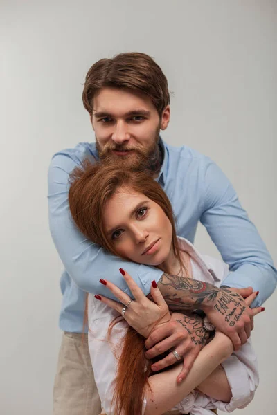 Beautiful fashionable young couple in stylish elegant casual clothes in the studio. Beautiful young happy redhead woman and handsome fashionable hipster man with tattoo and beard hug
