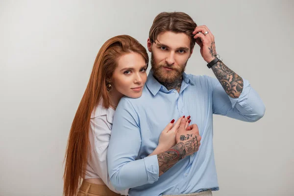 Beautiful fashionable young couple in elegant trendy clothes on a white background. A beautiful redhead young girl hugs a handsome brutal man with a beard and a tattoo fixes her hair