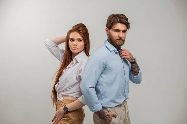 Stylish beautiful young redhead woman model in fashion clothes and handsome trendy hipster man with tattoo and beard in blue shirt