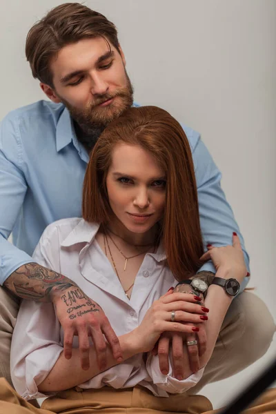 Handsome brutal man with a beard and a tattoo in a blue shirt hugs a beautiful red-haired woman in the studio. fashion couple
