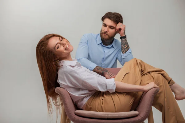 Happy beautiful young fresh redhead woman sitting in chair with smile and handsome brutal man with beard and tattoo on a white background
