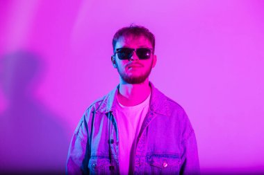 Modaya uygun güneş gözlüklü yakışıklı adam pembe arka planda pembe kot ceketli yaratıcı pembe ve mavi ışıklı.