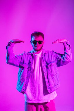 Moda modası kıyafetleri içinde, kot ceketli ve güneş gözlüklü yakışıklı hippi genç adam pembe ve mavi ışıkla yaratıcı stüdyoda dans ediyor.