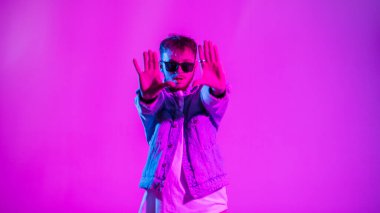 Moda kot ceketli, şık güneş gözlüklü yakışıklı hip hop dansçısı yaratıcı pembe ve mavi ışıkla renkli stüdyoda dans ediyor.