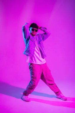 Profesyonel hip hop dansçısı, güneş gözlüklü, kot elbiseli, şapkalı adam pembe ve neon ışıklı yaratıcı bir stüdyoda dans ediyor.