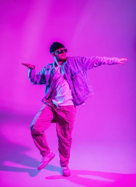 Kot ceketli, güneş gözlüklü, şapkalı ve spor ayakkabılı profesyonel dansçı stüdyoda yaratıcı pembe ve neon ışıklarla dans ediyor.
