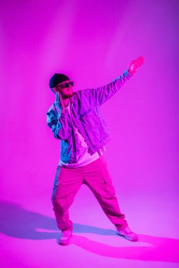 Moda gözlüklü ve şık kot elbiseli yakışıklı dansçı hip hop adam stüdyoda yaratıcı pembe ve mavi ışıkla dans ediyor.