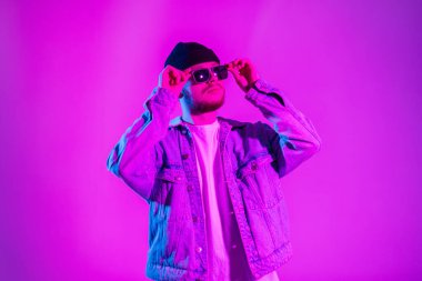Moda kot ceketli şık, yakışıklı hippi adam stüdyoda yaratıcı pembe ve mavi ışıklı güneş gözlüğü takıyor.