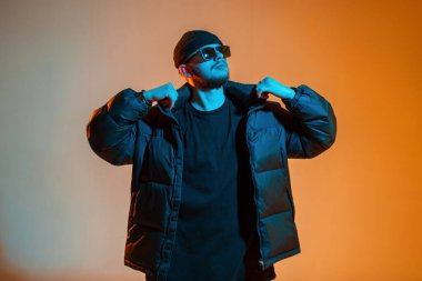 Şapkalı ve havalı güneş gözlüklü şık şık bir adam siyah modaya uygun ceketli yaratıcı renkli stüdyoda turuncu ve neon ışıklı pozlar veriyor.