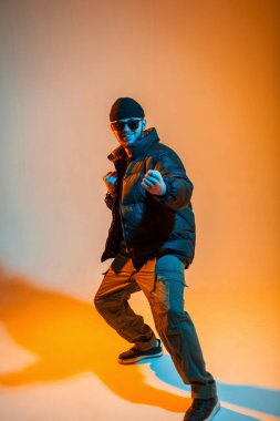 Şapkalı, güneş gözlüklü, modaya uygun siyah ceketli, yaratıcı turuncu ve neon ışıklı bir dansçı stüdyoda poz veriyor.