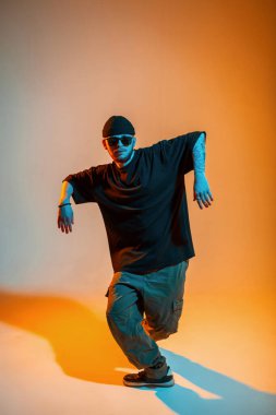 Moda kıyafetleriyle günbatımı ve şapkalı şık şık erkek dansçı. Yaratıcı bir stüdyoda turuncu ve neon ışıklarla dans ediyor.