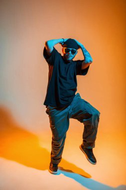 Siyah şapkalı, güneş gözlüklü, tişörtlü ve spor ayakkabılı yakışıklı dansçı turuncu bir arka planda dans ediyor.