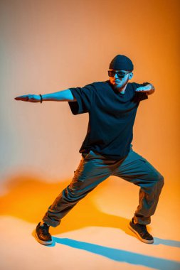 Şapkalı, güneş gözlüklü, tişörtlü ve spor ayakkabılı profesyonel hip hop dansçısı turuncu ve neon ışıklı bir stüdyoda dans ediyor.