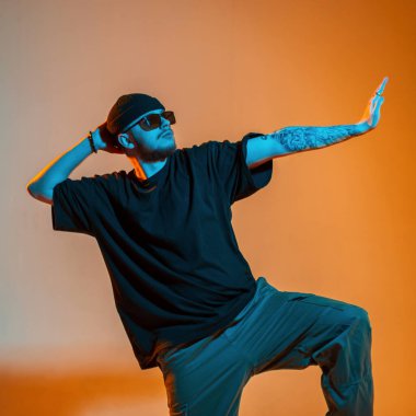 Siyah tişörtlü ve şapkalı yakışıklı profesyonel dansçı turuncu ve neon ışıklı yaratıcı bir stüdyoda dans ediyor.