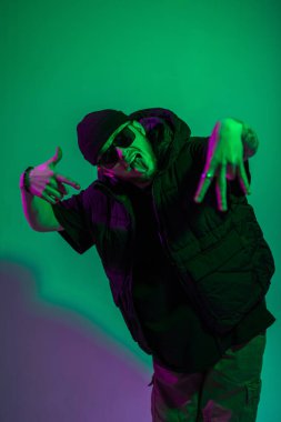 Moda gözlüklü havalı hippi adam ve siyah elbiseli bir şapka. Renkli bir stüdyoda yeşil ve mor ışıklı poz veriyor.