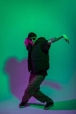 Havalı profesyonel hip hop dansçısı şapkalı ve güneş gözlüklü moda siyah elbiseli yaratıcı bir renk stüdyosunda yeşil ve pembe ışıklı dans ediyor.